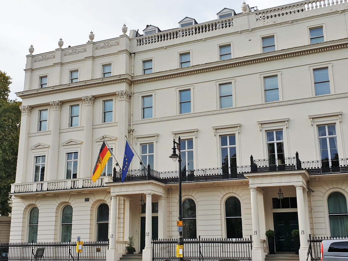 The pale stone front of an embassy building on a quiet London street, with national and EU flags above the entrance.