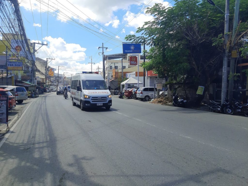A white ambulance van driving down a sunny city street lined with shops and power lines.