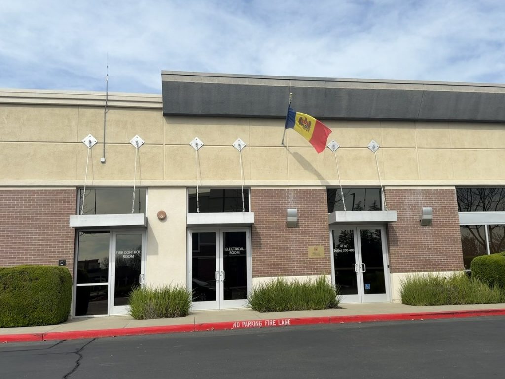 The entrance of a low brick consulate building with glass doors and a national flag mounted above them.