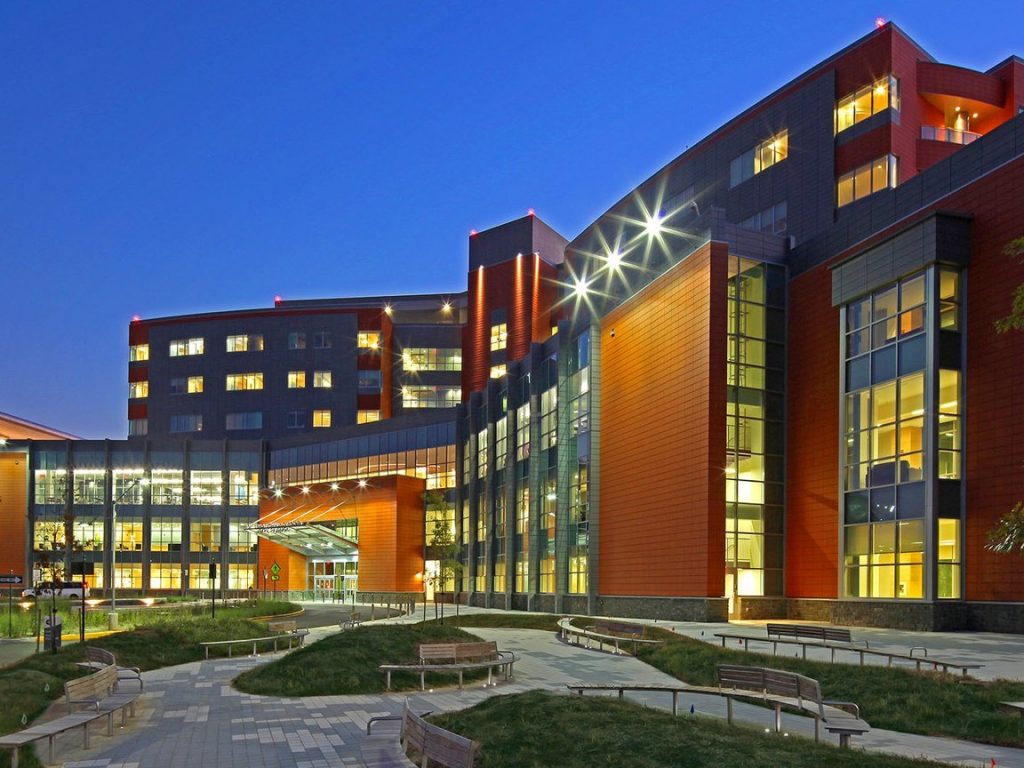 A modern hospital building with lit windows at dusk, with a landscaped walkway in front.