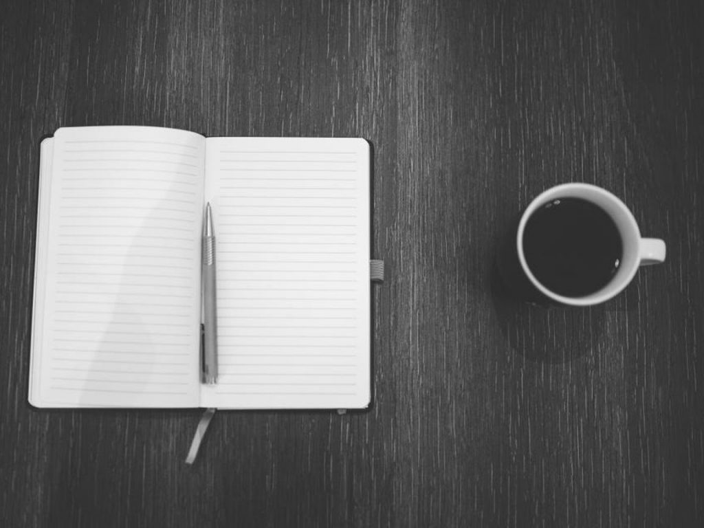 An open lined notebook with a pen beside a cup of black coffee on a dark table.