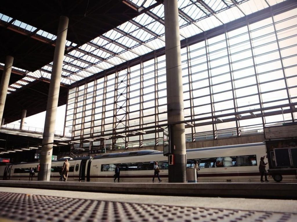 A large train station hall with a glass roof, steel columns, and a train waiting at the platform.