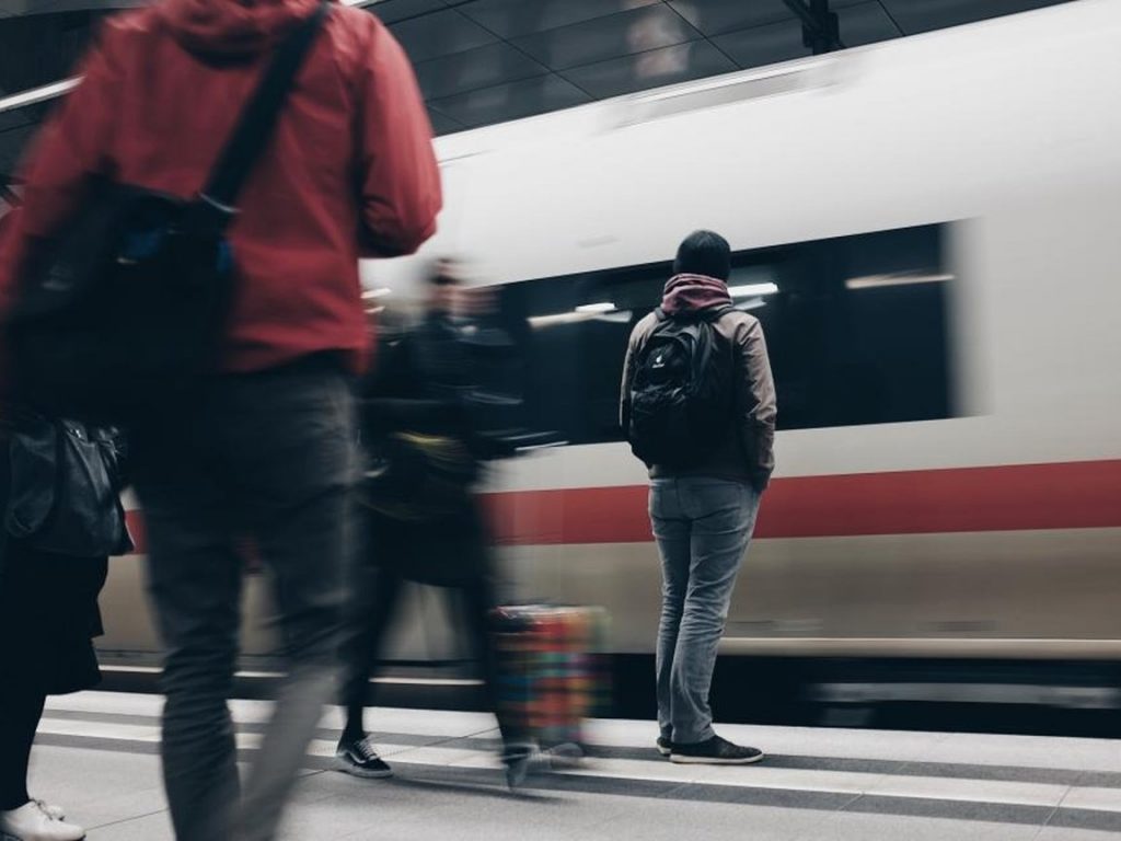 Travelers with backpacks walking along a station platform as a high-speed train passes, seen from behind.
