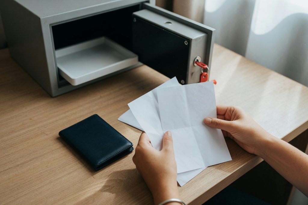 A traveler sorts plain photocopies beside a closed passport cover next to an open hotel room safe.