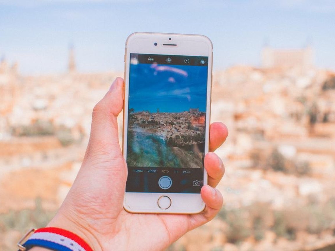 A hand holding a smartphone up against a blurred view of an old hilltop town.