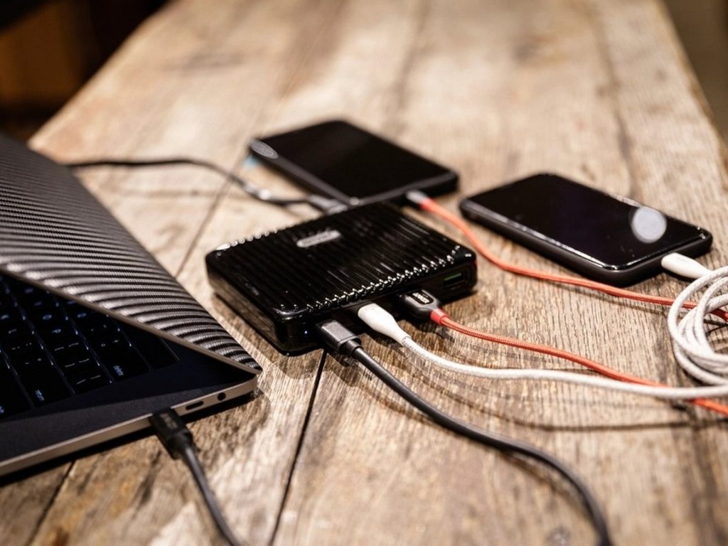 Two smartphones charging from a portable power bank on a wooden table next to a laptop.