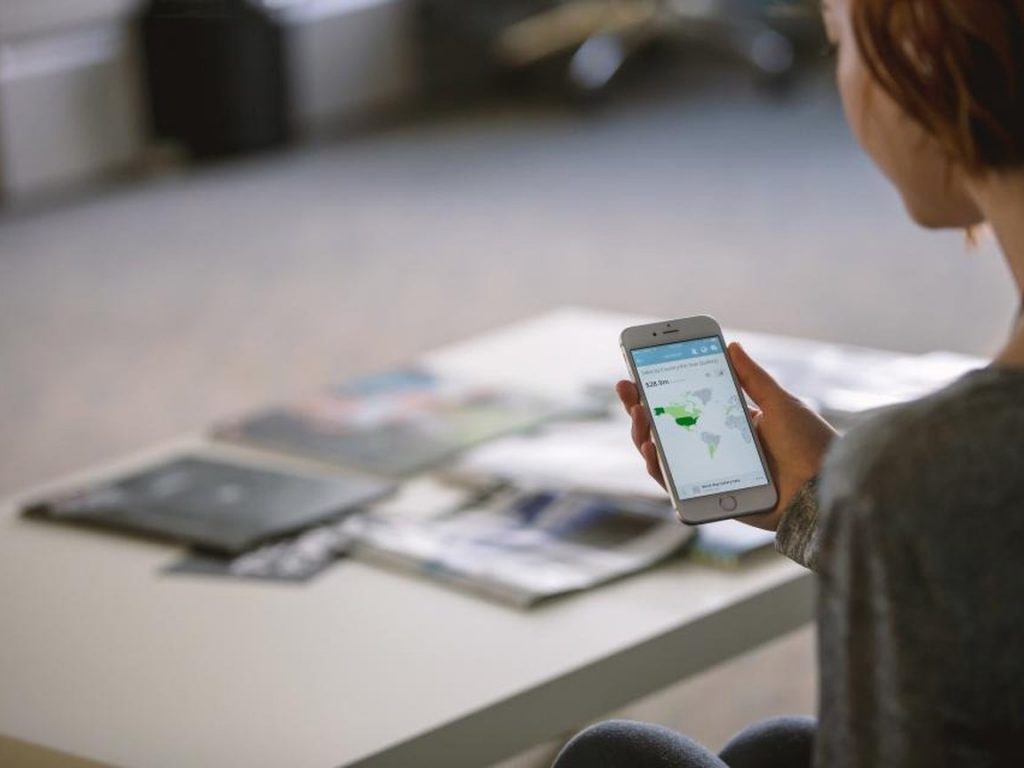 A person seated at a table checks a smartphone held in one hand, with travel brochures spread in front of them.