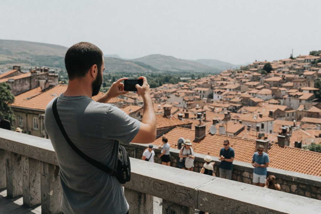 A bearded traveler photographs terracotta rooftops and distant hills from a busy public viewpoint at midday.