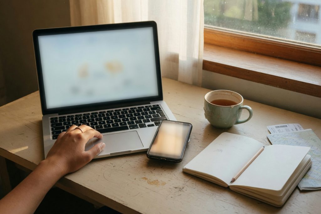 A laptop, a phone, a notebook, and a cup of tea on a wooden desk by a window as a traveler saves notes.